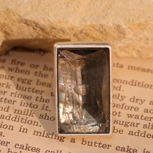 May include: A rectangular, smoky quartz gemstone set in a silver-colored bezel. The gemstone has a faceted cut, reflecting light. The setting is simple and elegant. The background is a vintage book page.