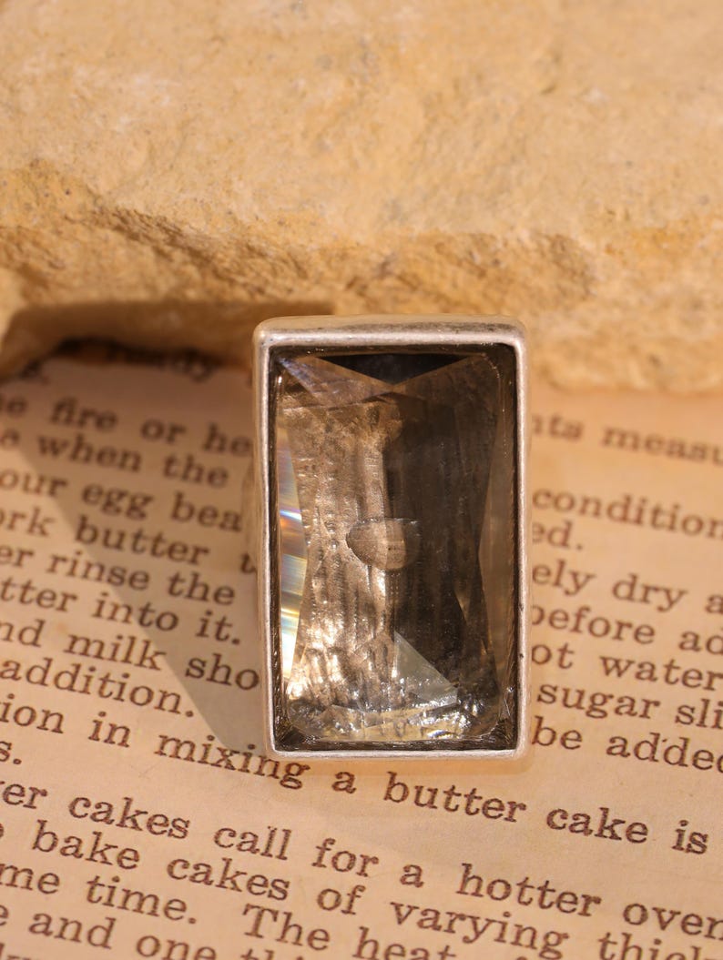 May include: A rectangular, smoky quartz gemstone set in a silver-colored bezel. The gemstone has a faceted cut, reflecting light. The setting is simple and elegant. The background is a vintage book page.