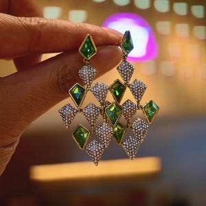 May include: Gold-toned chandelier earrings with green and clear gemstone accents. The earrings feature a geometric design with diamond-shaped green stones and smaller, clear stones. The earrings are held up by a hand.
