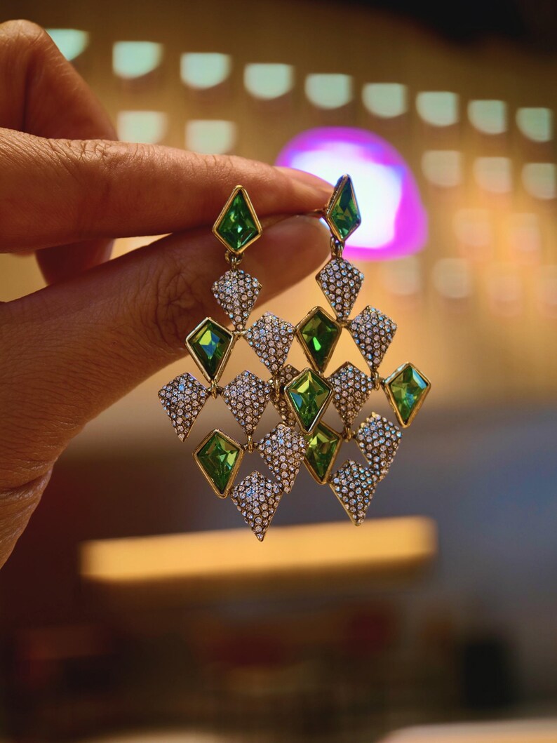May include: Gold-toned chandelier earrings with green and clear gemstone accents. The earrings feature a geometric design with diamond-shaped green stones and smaller, clear stones. The earrings are held up by a hand.