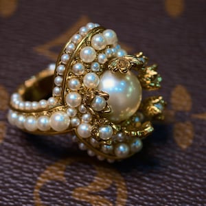 Puede incluir: Anillo dorado con una gran perla central rodeada de perlas más pequeñas. El anillo tiene un diseño detallado y ornamentado con una doble banda y detalles de perlas. Fondo marrón.