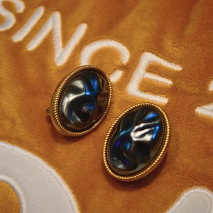 May include: A pair of oval-shaped earrings with a dark, reflective center stone, framed by a gold-colored, rope-like border. The earrings rest on a textured, orange fabric with the word "SINCE" embroidered in white.