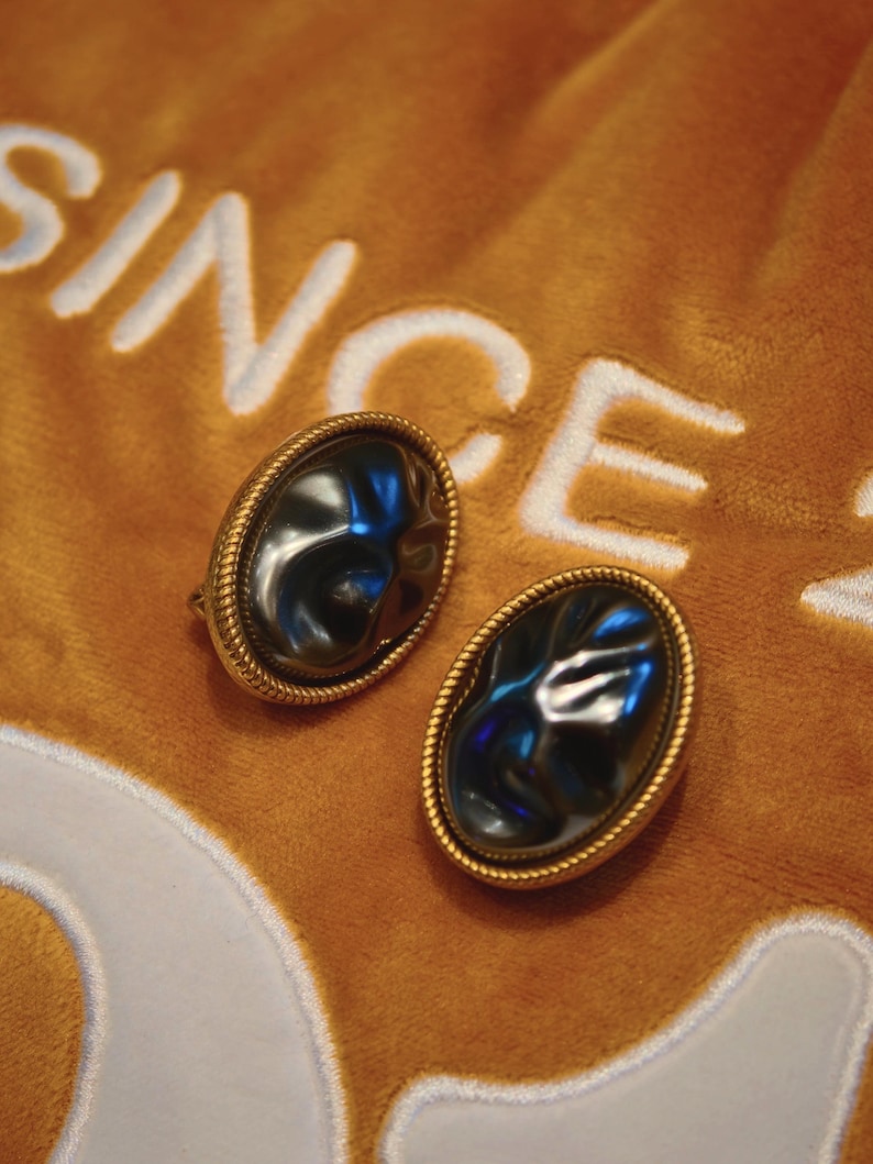 May include: A pair of oval-shaped earrings with a dark, reflective center stone, framed by a gold-colored, rope-like border. The earrings rest on a textured, orange fabric with the word "SINCE" embroidered in white.