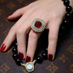 May include: A gold ring with a large, square, red gemstone, surrounded by smaller clear stones. A black beaded bracelet and a matching necklace with a gold clasp are also visible. The nails are painted a deep red.