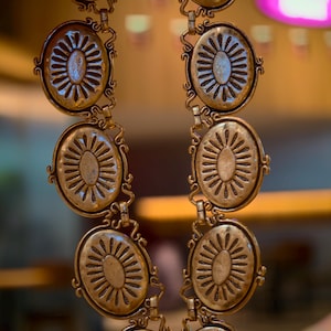 May include: A vintage-style gold-toned belt featuring oval medallions with a sunburst design. The belt is composed of multiple linked medallions, each with intricate detailing and a decorative border. The belt is held up by a hand.