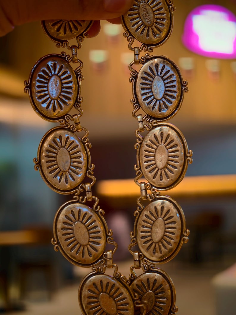 May include: A vintage-style gold-toned belt featuring oval medallions with a sunburst design. The belt is composed of multiple linked medallions, each with intricate detailing and a decorative border. The belt is held up by a hand.