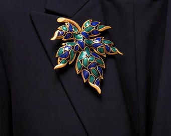 May include: A gold leaf brooch with blue and green enamel, pinned to a black blazer. The brooch is a detailed leaf design. The blazer has a Chanel label.