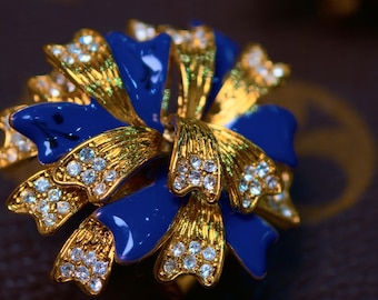 以下が含まれることがあります： 花型のジュエリーのクローズアップ。 金色の花びらにクリアなクリスタルのアクセントと鮮やかなブルーのエナメルが施されています。 デザインは複雑で人目を引き、高級アクセサリーを連想させます。