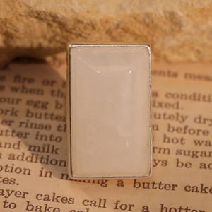 May include: A rectangular ring with a light cream-colored stone set in a silver-toned metal frame. The ring's design is simple and elegant, suitable for everyday wear. The ring is set on a vintage paper background.