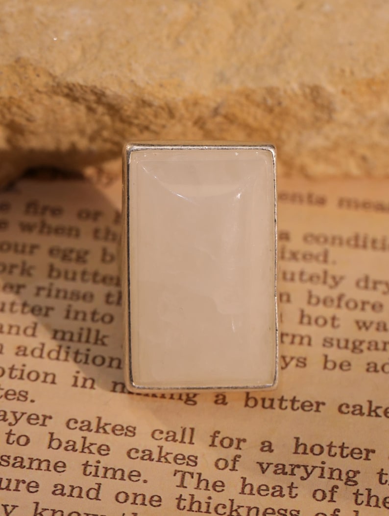 May include: A rectangular ring with a light cream-colored stone set in a silver-toned metal frame. The ring's design is simple and elegant, suitable for everyday wear. The ring is set on a vintage paper background.