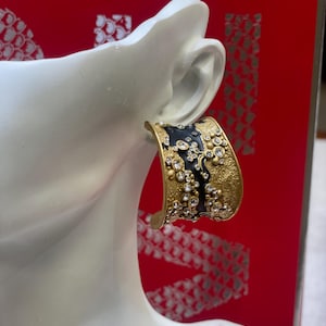 May include: A gold-toned cuff earring with a black enamel section and embellished with clear crystals. The earring is displayed on a white mannequin head against a red background.