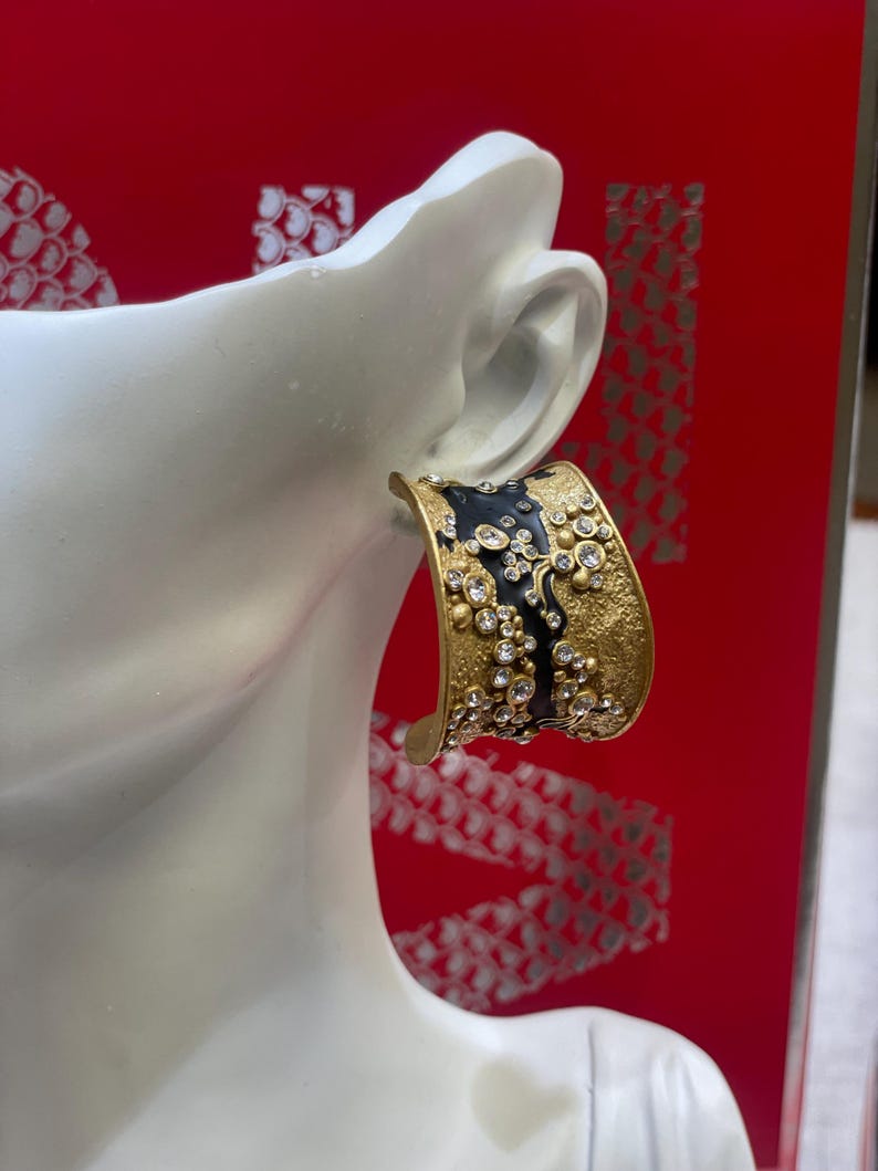 May include: A gold-toned cuff earring with a black enamel section and embellished with clear crystals. The earring is displayed on a white mannequin head against a red background.