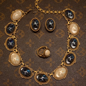 May include: A gold-toned jewelry set featuring a necklace, earrings, and a ring. The necklace and earrings have oval-shaped blue and white stones set in ornate frames. The ring has a similar design. The jewelry is displayed on a brown surface.