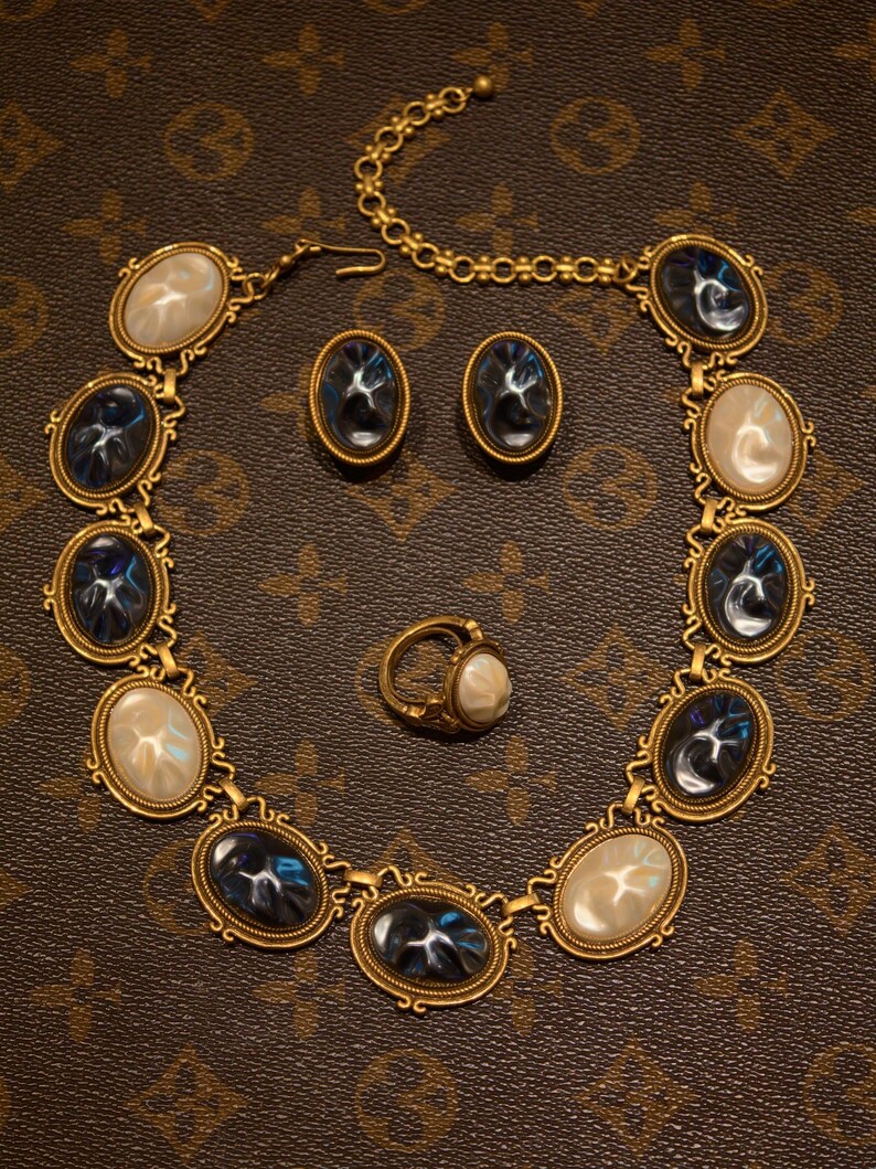 May include: A gold-toned jewelry set featuring a necklace, earrings, and a ring. The necklace and earrings have oval-shaped blue and white stones set in ornate frames. The ring has a similar design. The jewelry is displayed on a brown surface.