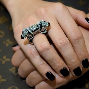 Puede incluir: Un anillo negro y plateado con una gran piedra central de corte cuadrado y gemas más pequeñas alrededor. El anillo presenta un diseño único con detalles intrincados y se muestra sobre un fondo neutro.