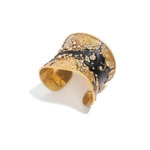 May include: A gold-toned cuff bracelet with a textured surface and black enamel accents. The bracelet is embellished with numerous clear, round gemstones. The design includes the text "GLOBAL JEWELRY".