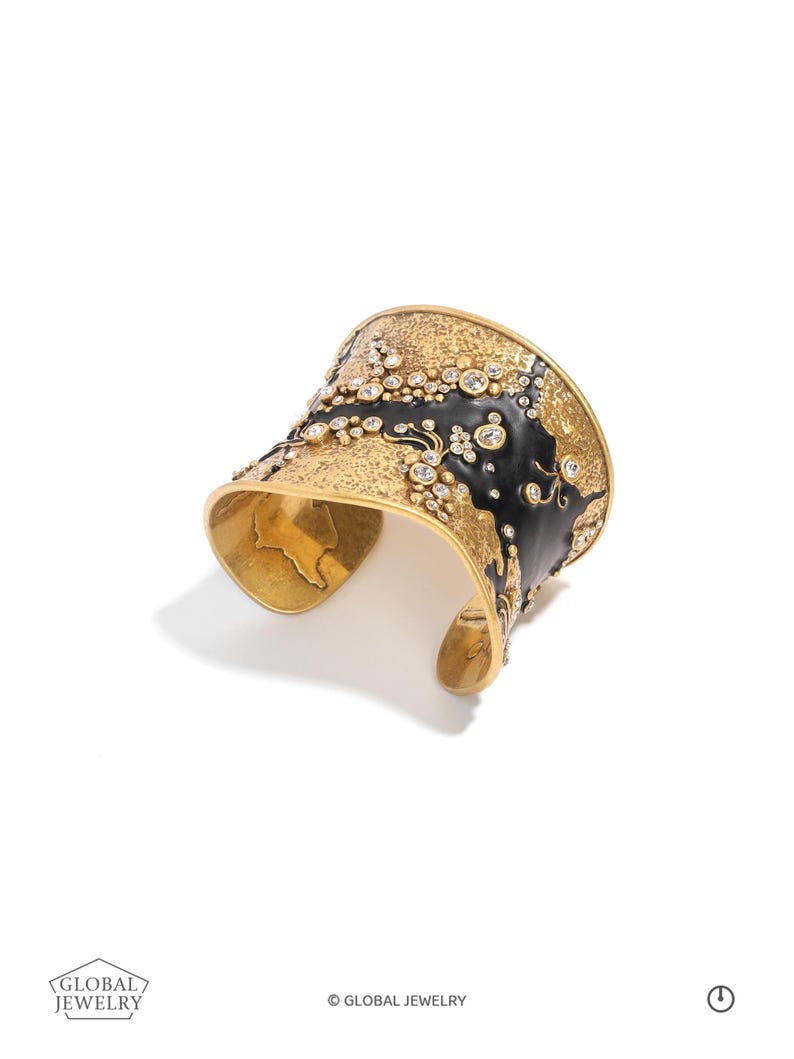 May include: A gold-toned cuff bracelet with a textured surface and black enamel accents. The bracelet is embellished with numerous clear, round gemstones. The design includes the text "GLOBAL JEWELRY".