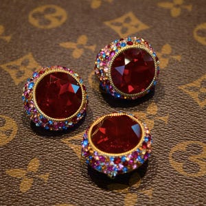 以下が含まれることがあります： 大きな赤い宝石が特徴の、装飾的で丸いイヤリングが3つ。各イヤリングは、ピンク、青、黄色の小さなカラフルなクリスタルバンドで囲まれ、金色の金属にセットされています。