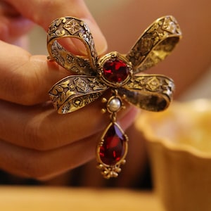 May include: A gold-coloured bow brooch with detailed floral patterns. It has a large, oval, red gemstone at the centre and a teardrop-shaped red gemstone pendant. A small pearl hangs below the central gemstone.