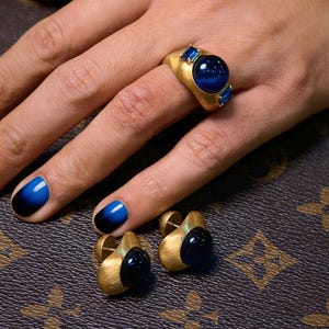 May include: A gold-toned ring with a large, round, blue gemstone and two smaller blue gemstones. Matching gold-toned earrings with blue gemstones are also present. The jewelry is displayed on a patterned surface.