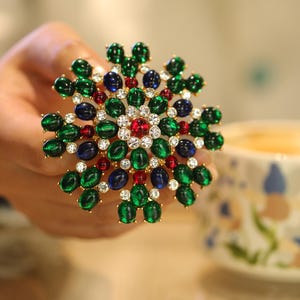 Peut inclure: Broche dorée avec un motif floral. Elle présente des pierres précieuses ovales vertes, bleues et rouges, ainsi que des cristaux transparents. Le centre de la broche est orné d'une pierre rouge entourée de cristaux.