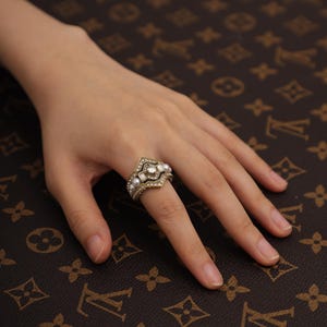 May include: A gold-toned ring featuring a central square-cut clear stone, surrounded by a geometric frame of smaller clear crystals and accented with small white pearls along the band, creating a layered and ornate appearance.