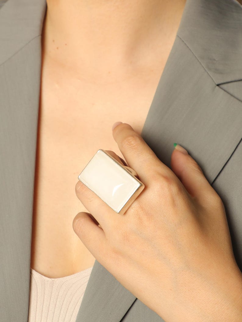 May include: A large, rectangular ring with a cream-colored, glossy surface. The ring's shape is a simple rectangle with a silver-colored band. The ring is worn on a finger, and the background includes a gray blazer and a hint of skin. Adjustable comfort fit — each ring contains a discreet inner mechanism that gently adapts to your finger size.
