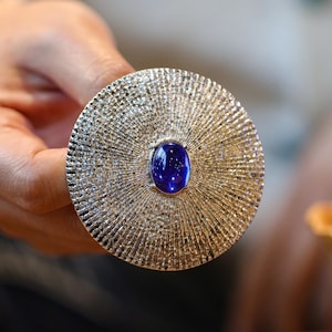 May include: A large, circular, gold-coloured jewellery piece with a textured, radial pattern. It has a vibrant, oval, deep blue gemstone at its centre. The piece is held by a hand, highlighting the intricate details and craftsmanship of the jewellery.