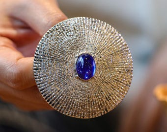 May include: A large, round, gold-toned jewelry piece with a textured, radial design. The center features a vibrant, oval-shaped, deep blue gemstone. The piece is held up by a hand, showcasing the intricate details and craftsmanship of the jewelry.