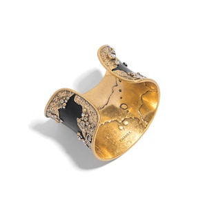 May include: A gold-toned cuff bracelet with black accents and sparkling embellishments. The interior has a textured finish. The bracelet has a curved design and is set against a white background. The word "CHAREL" is visible on the inside of the bracelet.