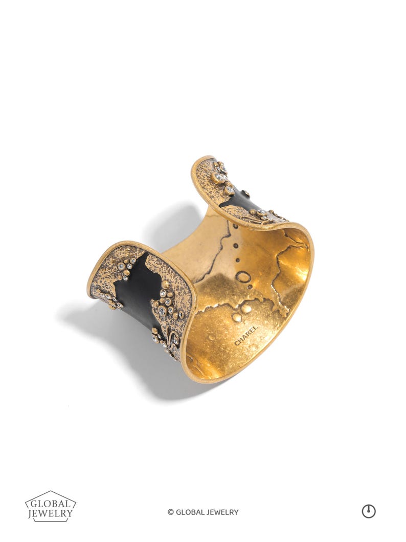 May include: A gold-toned cuff bracelet with black accents and sparkling embellishments. The interior has a textured finish. The bracelet has a curved design and is set against a white background. The word "CHAREL" is visible on the inside of the bracelet.