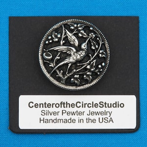 May include: A silver pewter jewellery pendant featuring a detailed hummingbird design with floral accents. The pendant is circular and has a black background. The text "CenteroftheCircleStudio Silver Pewter Jewellery Handmade in the USA" is printed on a white label below the pendant.