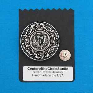 Könnte beinhalten: Silberner Zinn-Anhänger mit einem Distel-Design und filigranen Schnörkeln. Der Anhänger befindet sich auf einem schwarzen Hintergrund mit einem weißen Etikett, auf dem "CenteroftheCircleStudio Silber Zinn Schmuck Handgefertigt in den USA" steht.