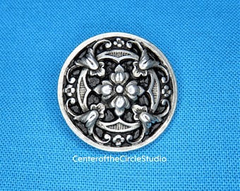 May include: A silver-toned button with a black center and intricate floral designs. The button has a raised, three-dimensional design.