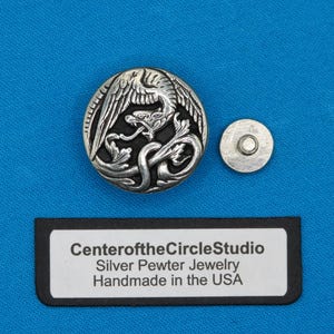 May include: A silver pewter button with an intricate design of a bird and a snake, next to a smaller silver button. The text "Center of the Circle Studio" is visible on a black label, along with "Silver Pewter Jewelry Handmade in the USA".