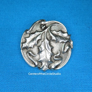 May include: A silver-toned metal pin featuring a detailed design of holly leaves.
