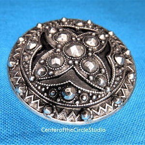 May include: A silver-toned metal button with a raised, intricate design. The button features a central motif of four overlapping, rounded shapes with a textured surface. The button is surrounded by a border of smaller, raised dots and a scalloped edge.