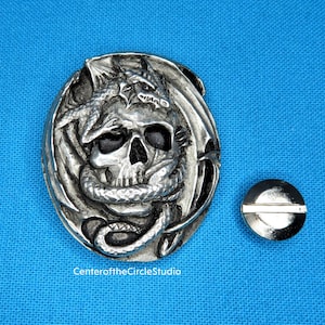 May include: A silver-toned metal belt buckle featuring a detailed design of a skull with a dragon wrapped around it. The buckle is circular and has a smooth, polished finish.