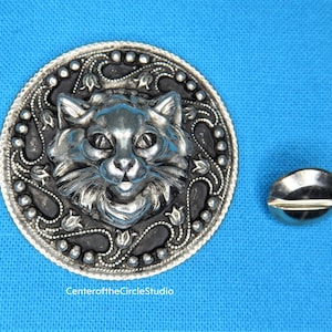 May include: A silver-toned metal brooch featuring a detailed cat head with a black background and a decorative floral border. The brooch is surrounded by a circle of small beads.