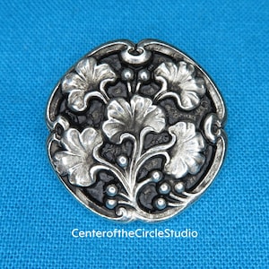 May include: A silver-toned button with a raised design of stylized leaves and berries. The button is round and has a black background.