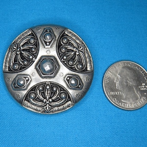 May include: A silver-toned, round, decorative button with an intricate design. The button is about the size of a US quarter.