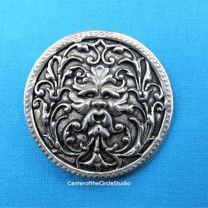 May include: A silver-toned metal button with an intricate design featuring a green man's face surrounded by swirling floral patterns.