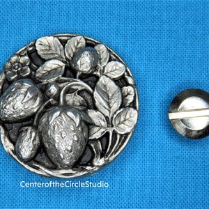 May include: A silver-toned metal button with a detailed design of strawberries and leaves. The button is round and has a black background. The button is about 2 inches in diameter.