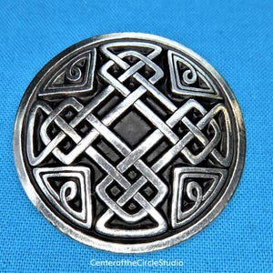 May include: A silver Celtic knot design brooch with a black background. The knot design is intricate and detailed, and the brooch is circular in shape.