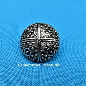 May include: A silver button with a decorative design of four circles with floral patterns. The button has a raised center and a smooth, shiny finish.