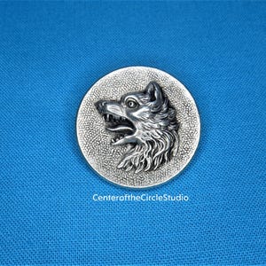 May include: A silver-toned metal pin featuring a detailed, three-dimensional wolf head design. The wolf's mouth is open, and its fur is textured. The pin is circular and has a textured background.
