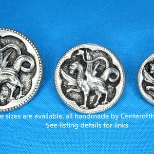 three silver-colored metal buttons with intricate designs, including winged creatures, against a blue background.