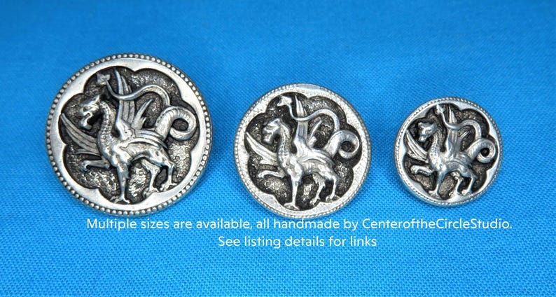 three silver-colored metal buttons with intricate designs, including winged creatures, against a blue background.