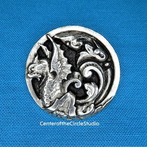 May include: A silver-toned button with a detailed design of a winged creature with a black background. The creature is in a curled position with its wings spread out. The button is round and has a smooth surface.