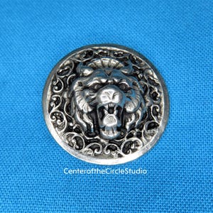 May include: A silver-toned button with a detailed design of a snarling lion's head in the center. The lion's head is surrounded by intricate floral patterns.